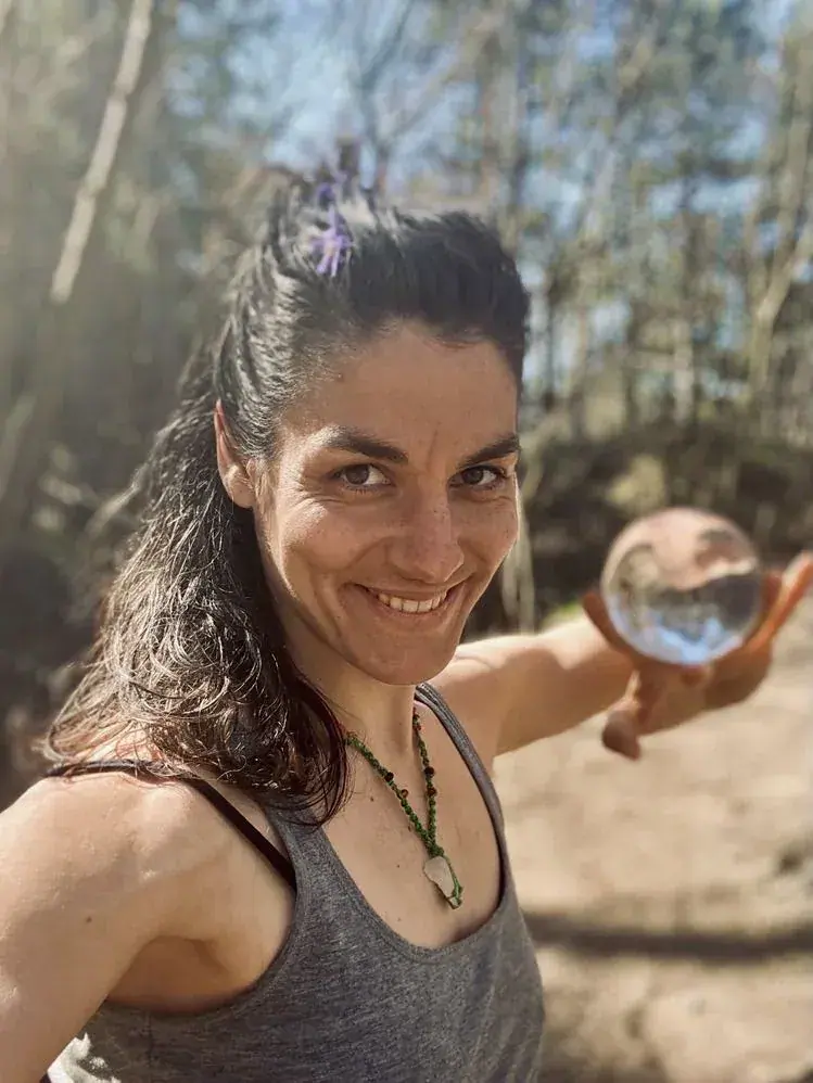 Smiling woman with a purple flower in her hair holds a clear crystal ball outdoors in a sunlit forest, for a Budapest