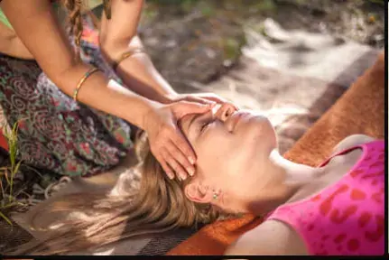 A masseuse gently placing her hands on the face of a woman lying on a mat outdoors, at Tantra Massage Paris