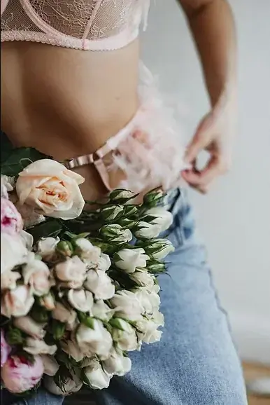 Cropped view of a woman in a pink lace bra and jeans holding a bouquet of cream and pink roses, Tantra Massage Paris