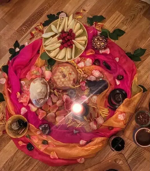 Overhead view of a ritual-style arrangement with a lit candle, sliced apples, berries and rose petals on colorful silk,