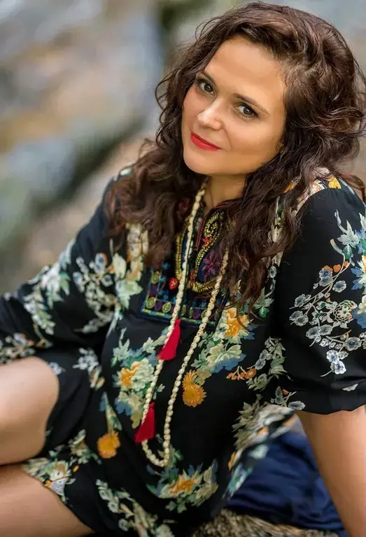 Woman with long curly dark hair in a black floral tunic and beaded tassel necklace, posing outdoors for Tantra Massage