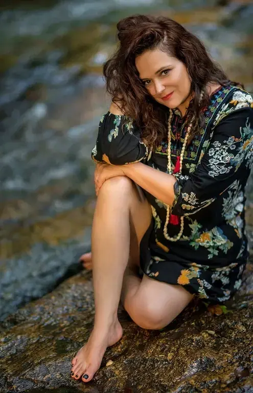 Smiling woman with dark wavy hair in a floral dress sitting barefoot on a rock by a stream, for Tantra Massage service in