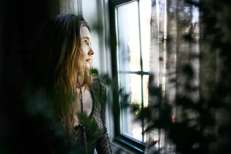 A woman with long hair gazes out a sunlit window in a moody, softly lit interior, used for Tantric Massage New York
