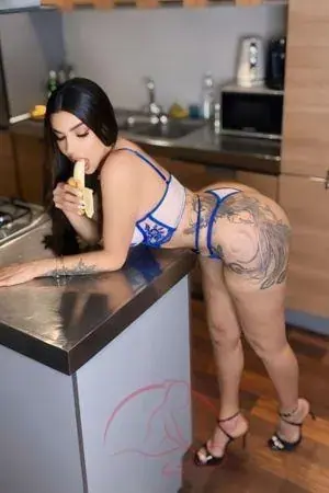 A woman in blue lace lingerie and heels leans on a kitchen counter eating a banana, promoting Tantric Touch in London